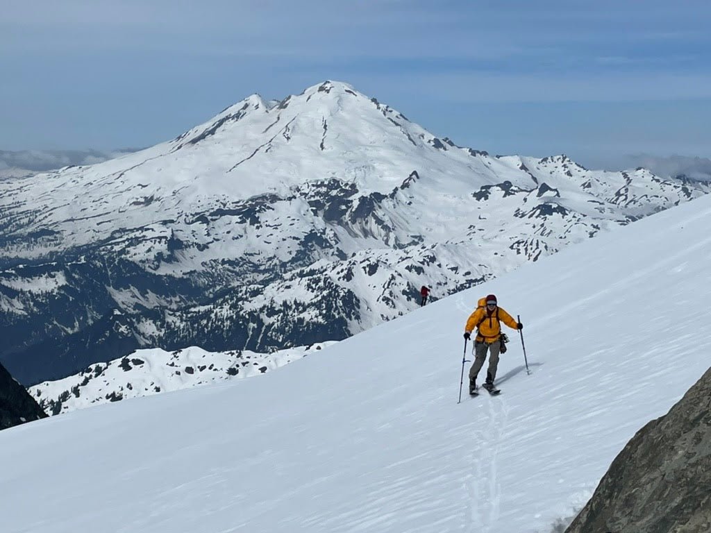 A splitboarder skins on Mount Shuksan Sulphide Glacier with Mount Baker in the background.