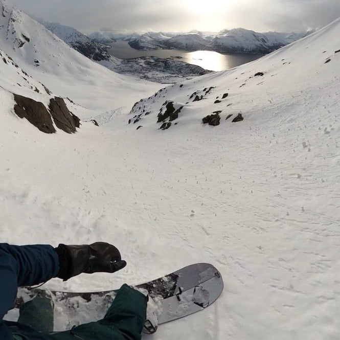 splitboarder rides a sunny slope through a couoir of rocks with the sun reflecting off ullsfjorden in northern norway