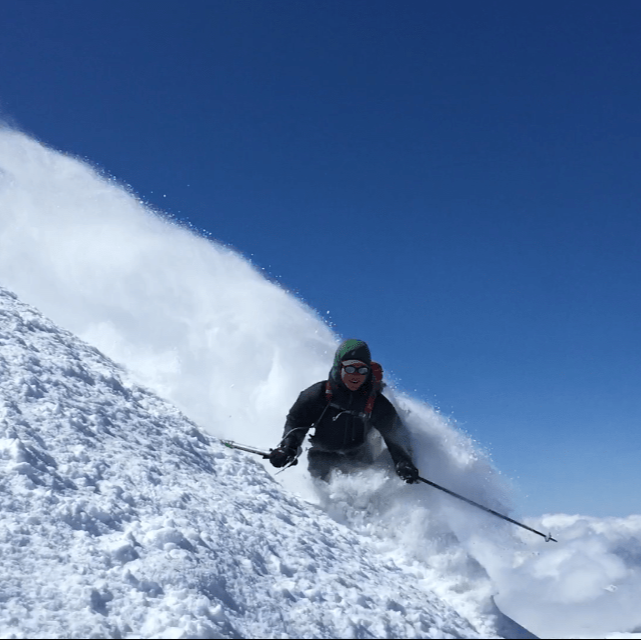 a skier makes a powder turn on mout shuksan whith blue skies behind them during a guided ski trip with guided exposure