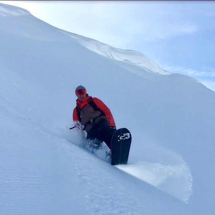 Stoked snowboarder riding in deep snow with pristine slopes and small cornices in the background.