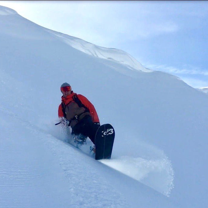 Stoked snowboarder riding in deep snow with pristine slopes and small cornices in the background.