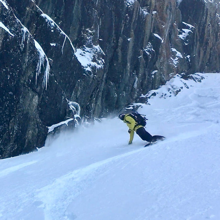 Snowboarder making turns on a snow covered slope next to a dramatic rock feature covered with snow and ice.