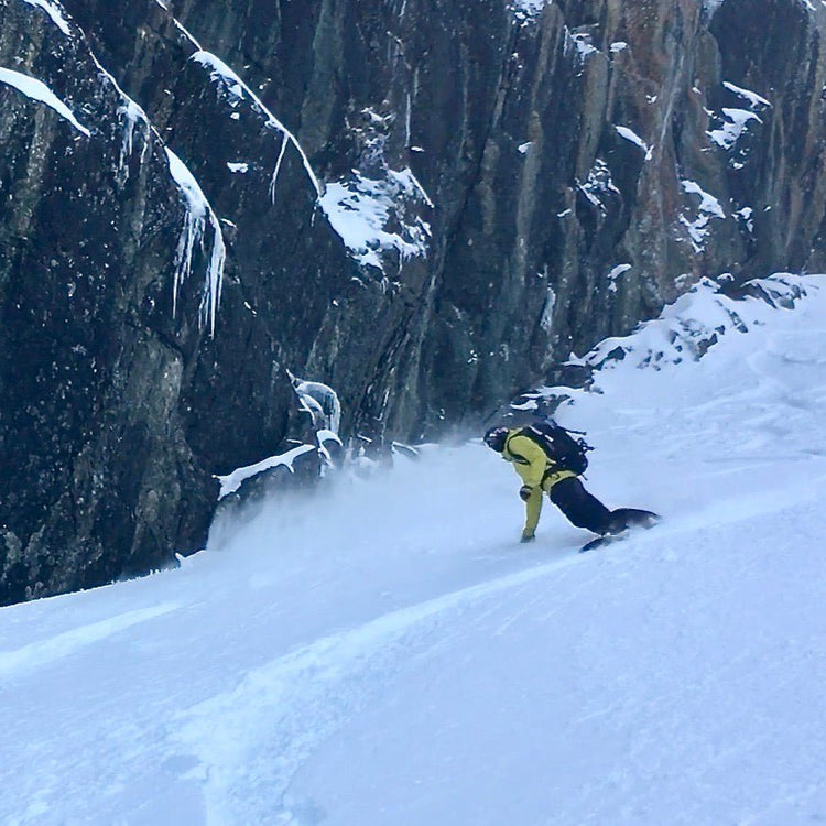 Snowboarder making turns on a snow covered slope next to a dramatic rock feature covered with snow and ice.