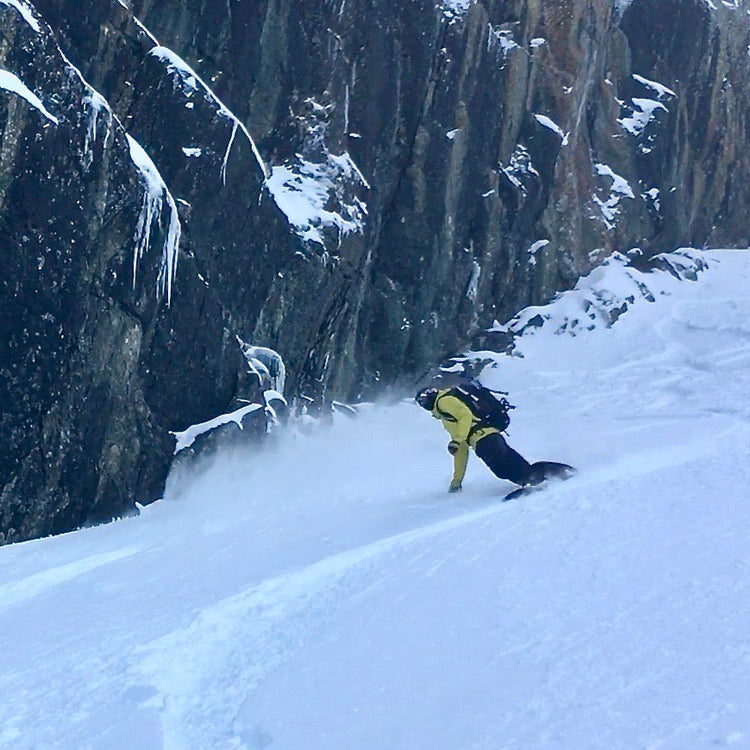 Snowboarder making turns on a snow covered slope next to a dramatic rock feature covered with snow and ice.