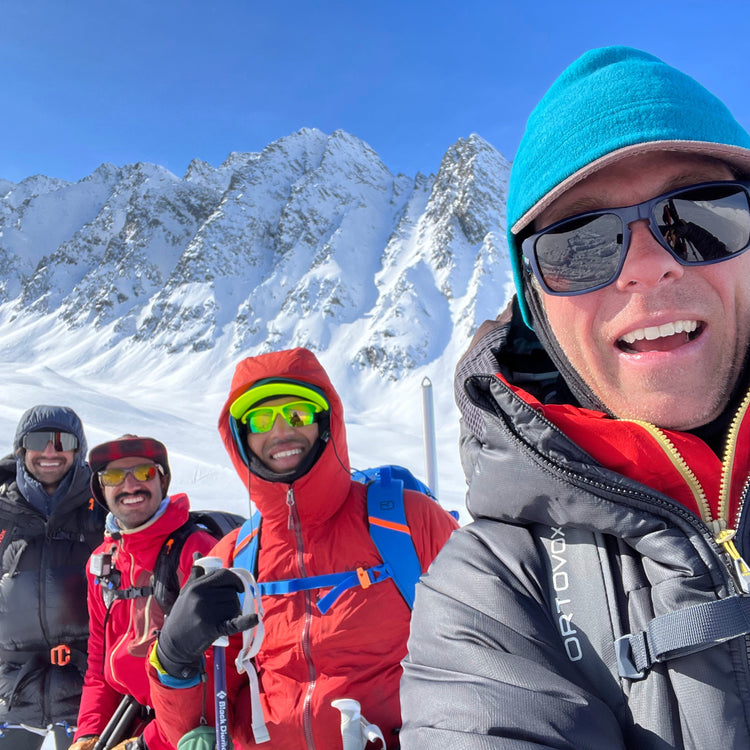 Four ski mountaineers smiling in front of steep, snow-filled couloirs under sunshine at Thompson Pass, Stairway Chutes, Valdez, AK