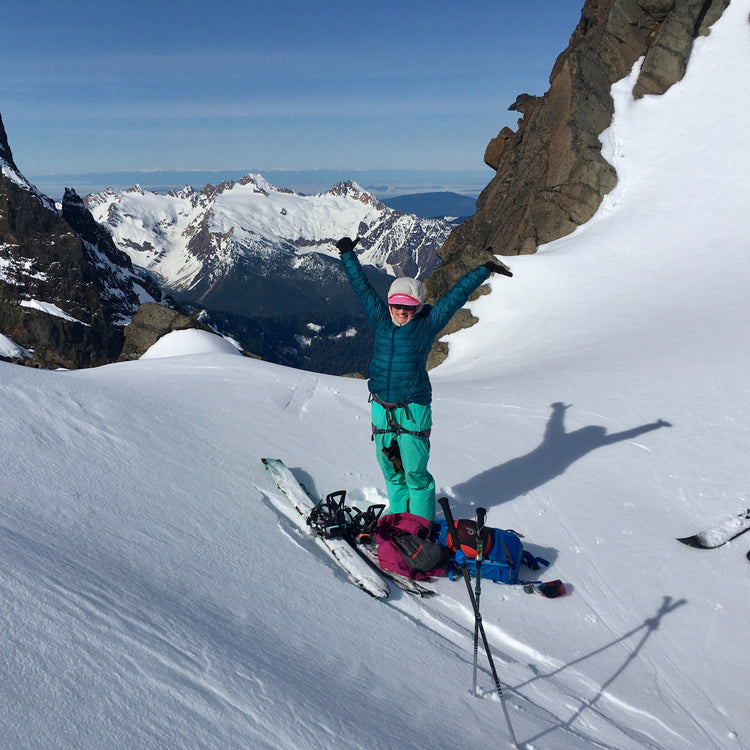a womens splitboard er cheers with hands in the air with snow and rocks framing the twin sisters range and olympic peninsula above bellingham washington