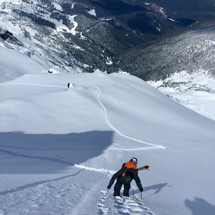 Looking back at a guest bootpacking up a steep snowy slope on the white salmon glacier mount shuksan, with a background of snow covered mountains. and the mt baker ski area