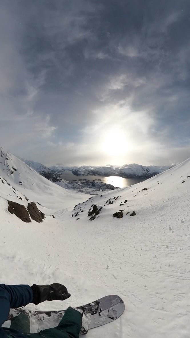 splitboarder rides a sunny slope through a couoir of rocks with the sun reflecting off ullsfjorden in northern norway