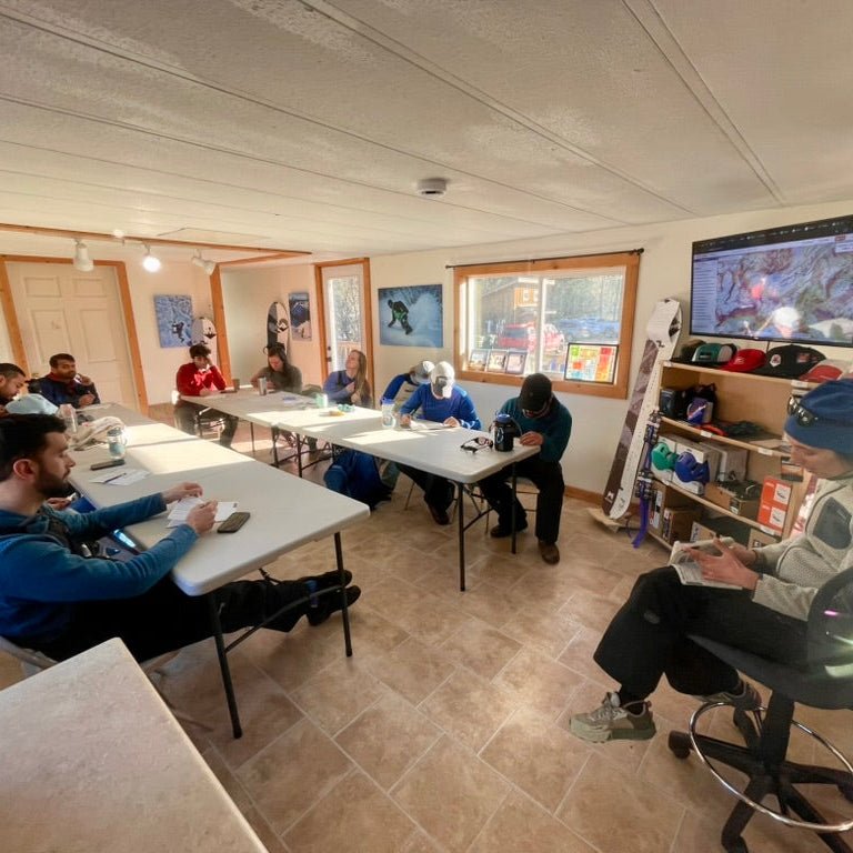 An AIARE avalanche course sitting preparing for the day in our Glacier WA classroom-like indoor space with windows, shelves, and a mounted screen on the wall.