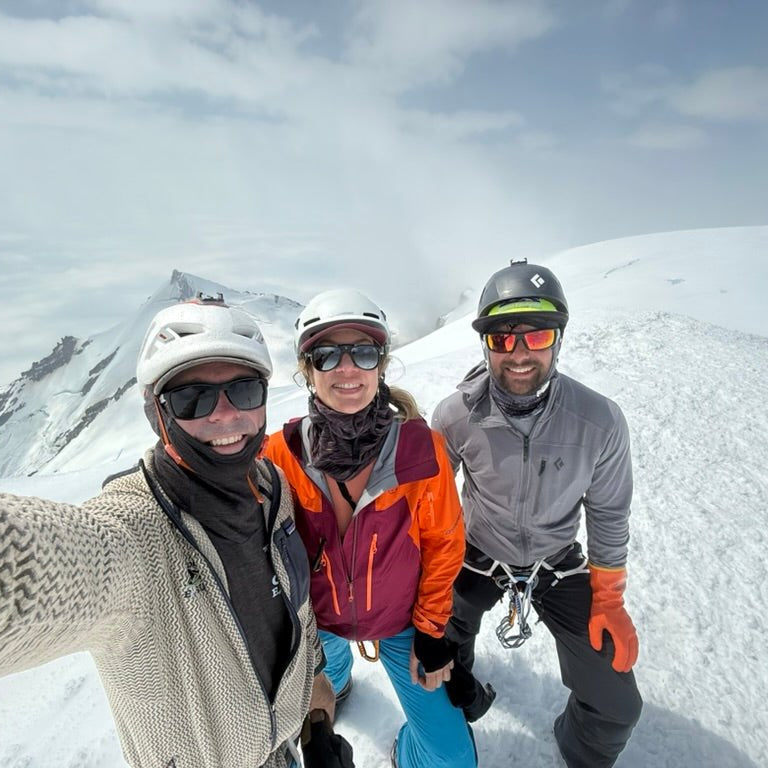 Three people posing for a selfie on a snowy mountain with ski equipment.