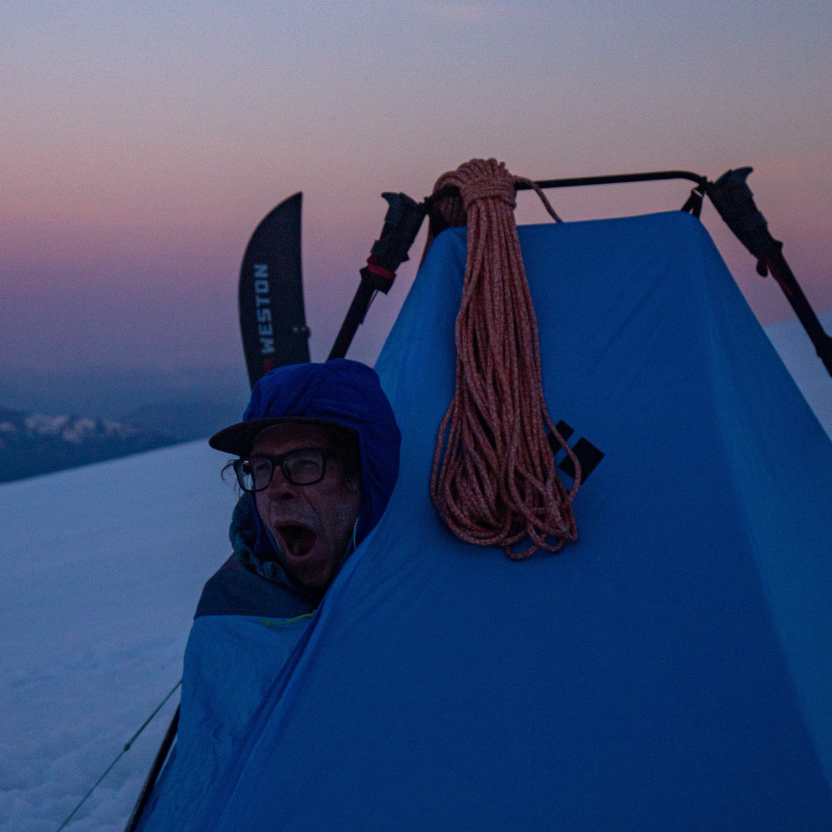 Person peeking out from a blue tent on a snowy mountain at dusk alpenglow of boulder glacier mount baker ski descent