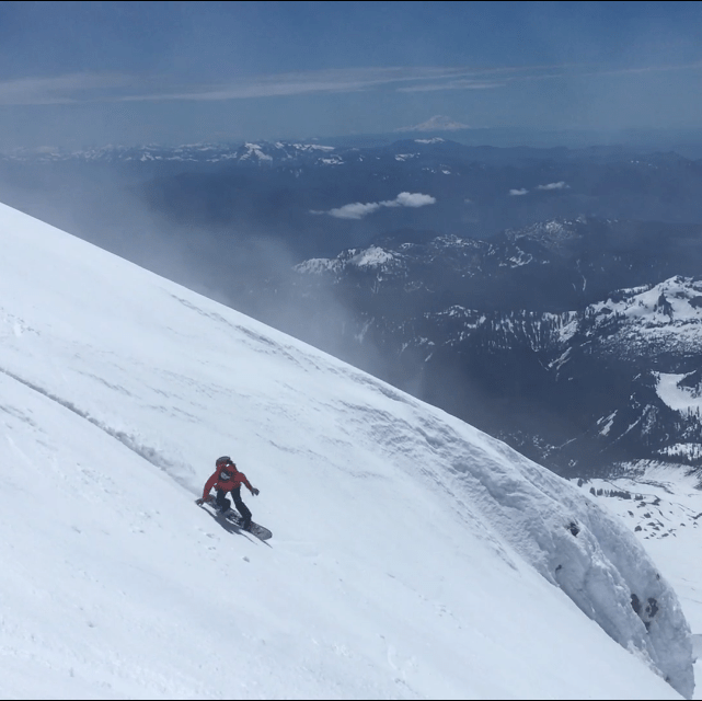 Person snowboarding down a snowy mountain with a vast mountain range in the background