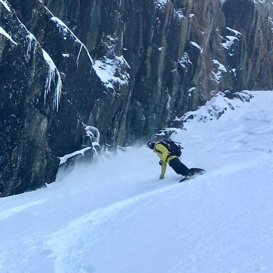 Snowboarder making turns on a snow covered slope next to a dramatic rock feature covered with snow and ice.