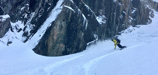 Snowboarder making turns on a snow covered slope next to a dramatic rock feature covered with snow and ice.