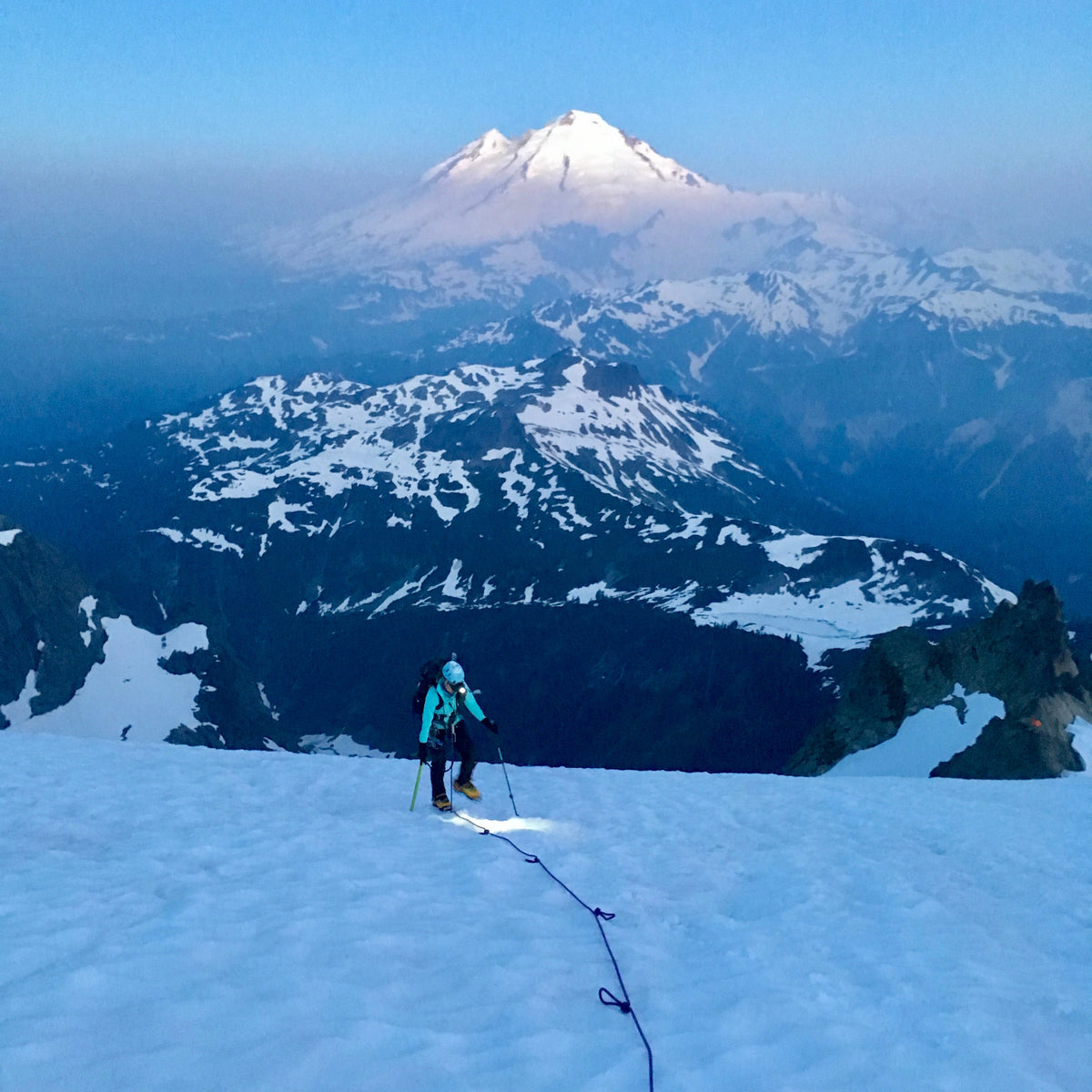 Walking up the curtis glacier on mount shuksan early in the morning, a light shadow of fog cloaks the lower mountains in the background, but Mount Baker rises above it. 