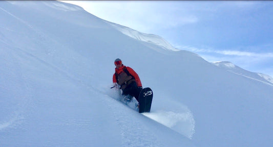 Stoked snowboarder riding in deep snow with pristine slopes and small cornices in the background.