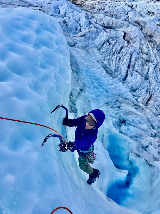 Ice climber swinging tools as they climb above a broken blue glacier.