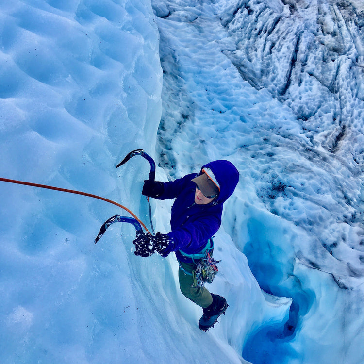 Ice climber swinging tools as they climb above a broken blue glacier.