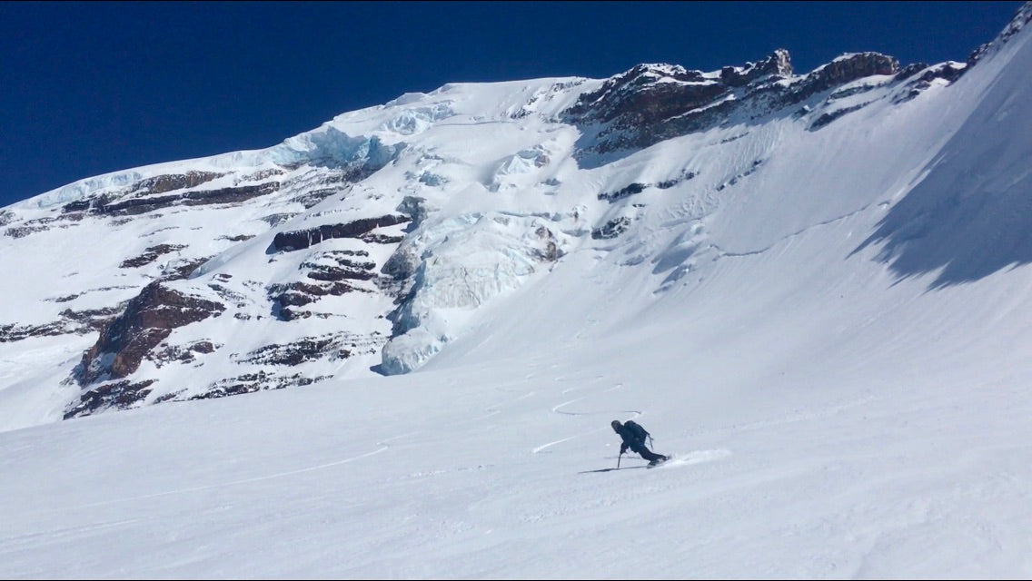 Splitboarder enjoying some low angle turns after riding steeps on Mount Baker. The mountain, covered in glaciers and snow, towers behind. 